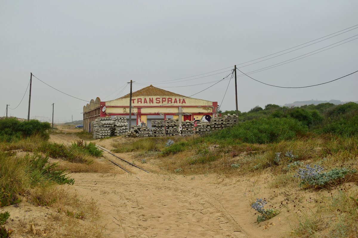 Blick auf die Strecke des Minicomboio da Caparica zwischen den Haltestellen Rainha und Praia da Rivieria auf das ehemalige Depot. Die Bahnstrecke wurde zwischen 1960 und 1962 als touristische Attraktion erbaut. 2008 wurde das erste mal über eine Stilllegung diskutiert die jedoch abgewendet werden konnte. Zu Beginn von Corona wurde der Betrieb eingestellt und bis heute nicht wieder aufgenommen. Es gibt jedoch gute Hoffnung das bald wieder Bahnen am Strand entlang fahren, denn die Strecke samt Fuhrpark wurde an jemanden verkauft der den Betrieb wieder herstellen möchte. 

Costa da Caparica 28.06.2024