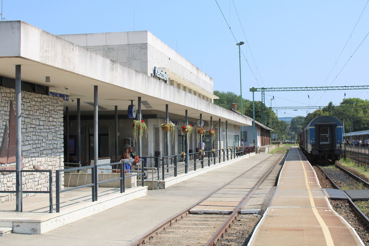 Das sehr gepflegte Bahnhofsgebäude mit dem schmalen Bahnsteig 1 und 2 am 19.07.2024 in Keszthely. Das Gleis 1 ist zudem mit Gleistragplatten aus Beton versehen. 