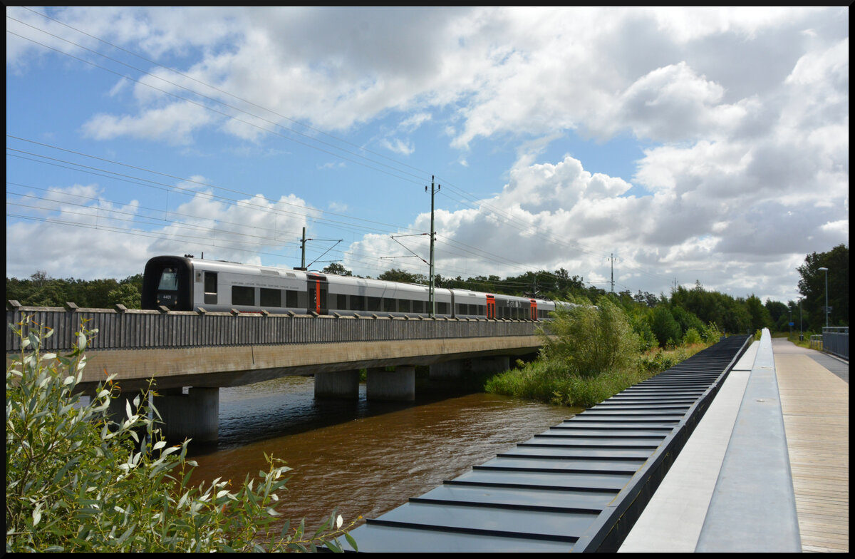 Der Öresundståg X31K/ET 4401 ist am 11.08.2024 auf dem Weg von Kopenhagen nach Göteborg, als er in Ängelholm diese moderne Brücke über den Fluss Rönne passiert. Die Aufnahme wurde von der alten Bahnbrücke aus aufgenommen. Sie dient heute Fußgängern und Radfahrern. 