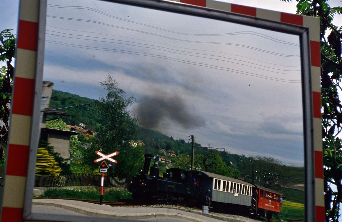 Schweizer Dampflok LEB G 3/3 Nr.5 mit einem kleinen Zug auf einer Spiegelung eines Schildes (der erste Wagen war ein Wagen der Montreux Oberland Bahn, der zweite Wagen zählte zur früheren Schweizer Privatbahn Chemins de fer electriques Veveysans). Der Zug war auf der Museumsbahn nach Chamby unterwegs (der Zug ist spiegelverkehrt auf dem Schild).
Datum: 18.05.1986