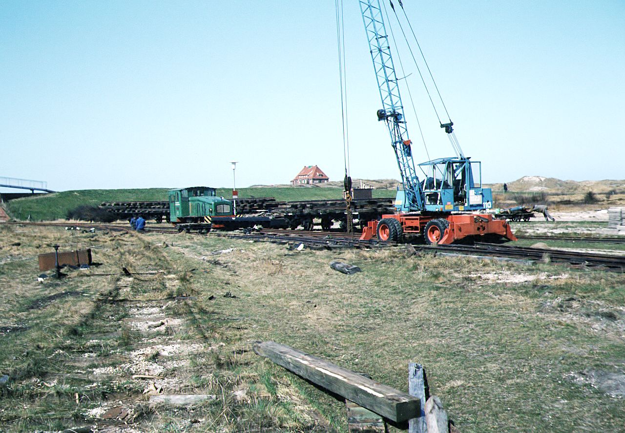 Inselbahn Spiekeroog__April 1984. Das ehemalige Bahnhofsgelände ist schon weitgehend  entgleist . Der Autokran verlädt Weichen und Gleisroste auf die Flachwagen zum Abtransport.