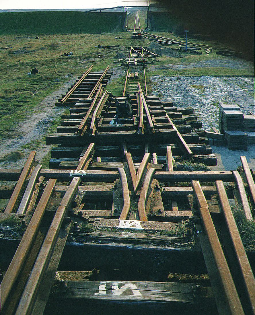 Inselbahn Spiekeroog__April 1984. Sauber nummeriert liegen Weichen-Teile für den Abtransport bereit. Der Blick geht im Abendlicht durchs geöffnete Deichtor auf die Strecke Richtung Westend.