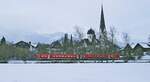 Bei leichtem Schneegriesel fährt ein 612 am 26.02.2023 an der Fischener Pfarrkirche St. Verena und der Frauenkapelle vorbei nach Oberstdorf