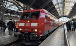 Der Coca-Cola Weihnachtszug steht im Leipziger Bahnhof. Auf der Vorderseite der rot-weißen Lok befindet sich das dB-Logo, während an der Seite der Lok der bekannte Coca-Cola-Schriftzug zu sehen ist. Der Zug ist im Zentrum des Bahnhofs abgebildet, umgeben von der Architektur des Gebäudes.