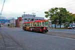 Strassenbahn Hakodate, im Süden der Insel Hokkaidô in Japan, Wagenserie 711-724: Wagen 718 fährt am Denkmal vorbei, welches an die Ankunft riesiger Zahlen von Geflüchteten erinnert, nachdem diese Menschen 1945 aus Sachalin vertrieben wurden. Bild vom 2.Oktober 2014  