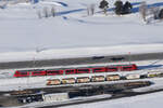 Nochmal RhB Rot am 17.01.2025 in der Früh zwischen Samedan und Punt Muragl vom Berg Muottas Muargl aus fotografieret. Der Zug passiert den Güterbahnhof bei Samedan.