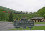 Eine der berühmtesten Eisenbahnbauwerke der USA ist die Hufeisenkurve (Horseshoe Curve) in der Nähe von Altoona, Pennsylvania. Sie ist Teil der Strecke von Harrisburg nach Pittsburgh über die Allegheny Mountain. Für Besucher hat man einen kleinen Park angelegt, von wo aus man die schwer arbeitenden Maschinen gut erleben kann. Leider ist es aber so gut wie unmöglich einen Zug in der Kurve komplett in Szene zu setzen. Hier bleibt dann nur des live Erlebnis zu bewahren, wenn man von einem Zug fast komplett umrundet wird, wo die Zugloks schon verschwunden sind und die Schubloks noch nicht zu sehen sind. Wer in der Gegend von Altoona ist, der sollte sich einen Ausflug zur Horseshoe Curve nicht entgehen lassen. Wenn man vorher das 'Railroaders Memorial Museum' in Altoona besucht hat und das Besucherbändchen noch trägt muß man auch kein zweites mal Eintritt zahlen!
Auf dem Bild, welches am 19. Mai 2018 entstand sieht man über den Hinweisschild die talwärts fahrenden Norfolk Southern Maschinen 9463 (GE C40-9W) und 9912 (GE C40-9W).